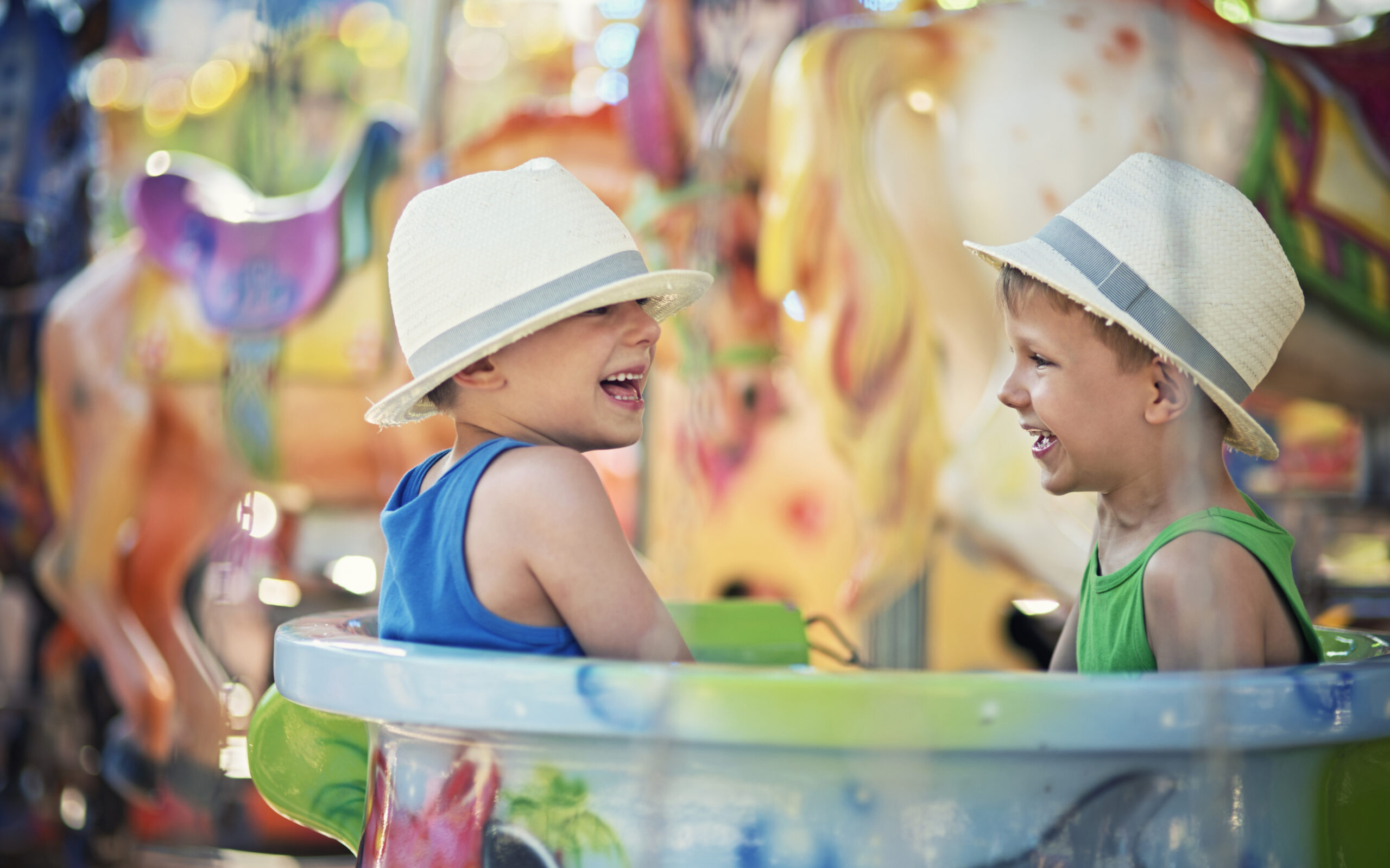 Two little brothers sitting in the same cup on carousel. The boys are aged 5 and wearing fedoras. Boys are having fun and laughing.